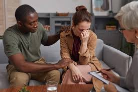 A couple in a session, one partner comforting the other while a therapist listens