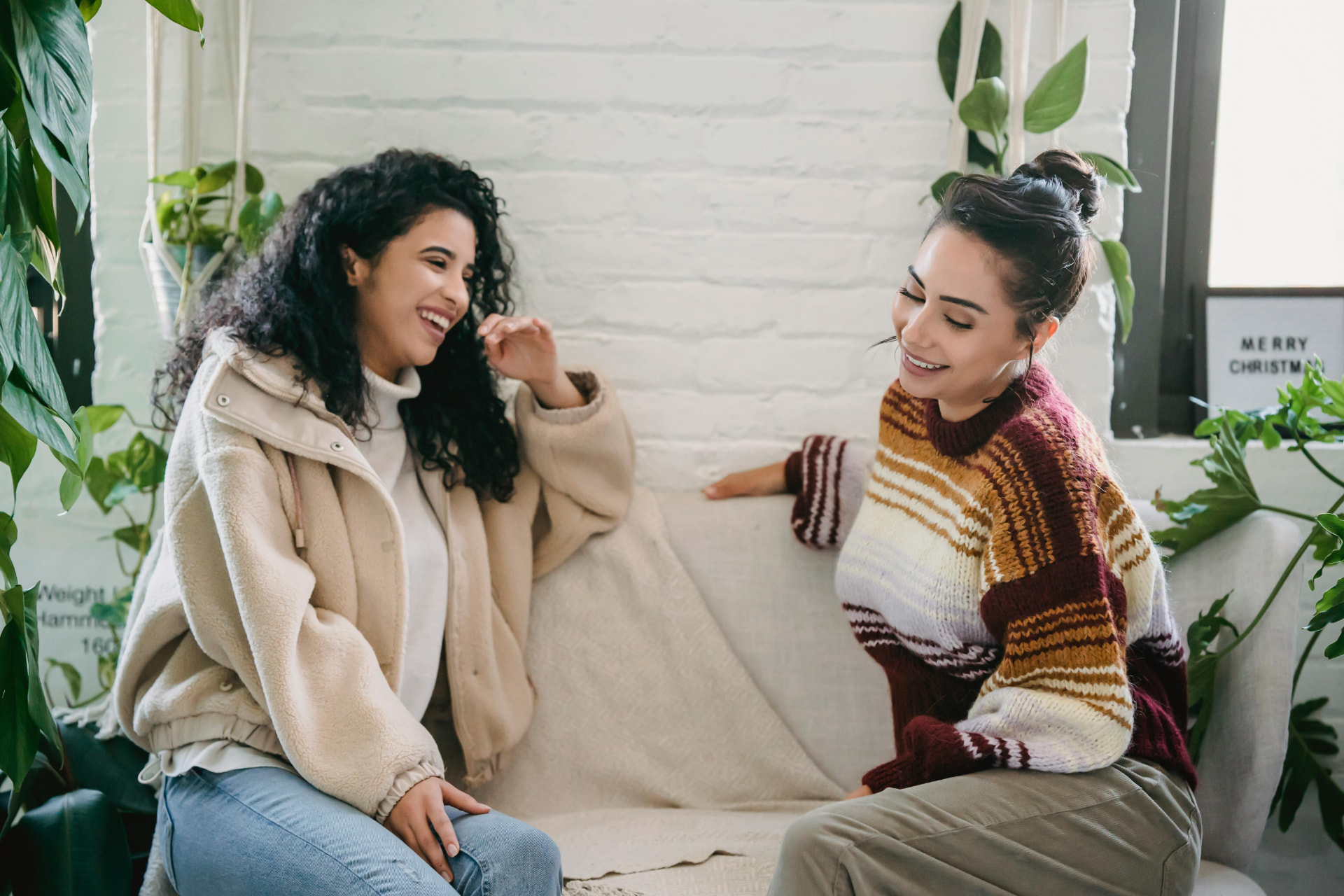Two women laughing together on a couch in a bright, plant-filled room